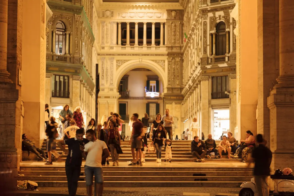 Crowd milling on broad marble steps inside ornate historic arcade at night.