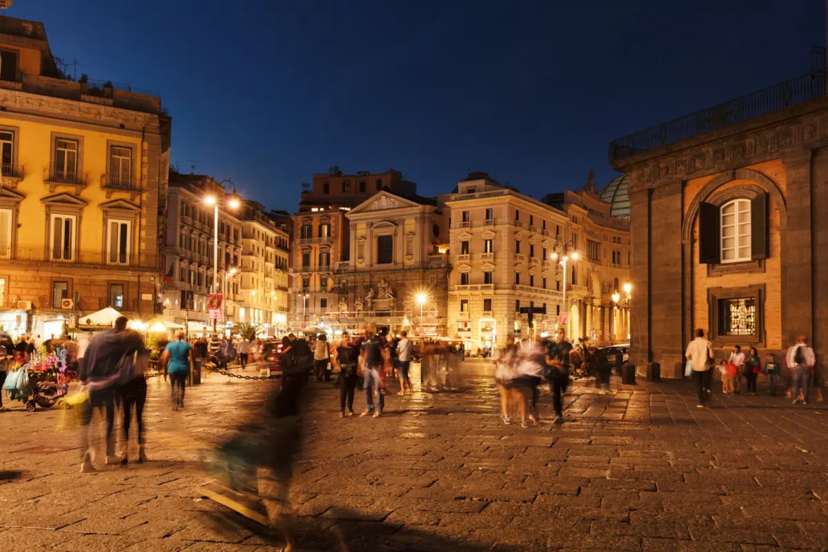 Pedestrians strolling in a busy evening plaza framed by warmly lit historic buildings and street stalls.