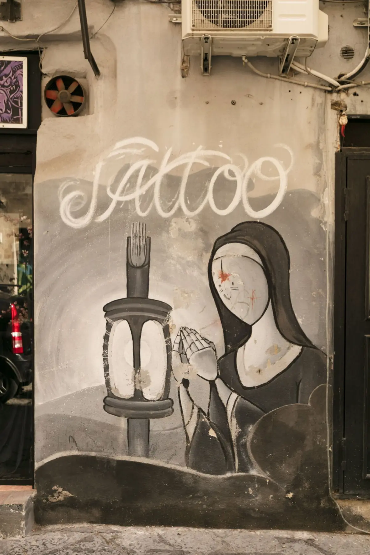 Hooded figure praying beside a tall candle in a graffiti mural on a worn wall.