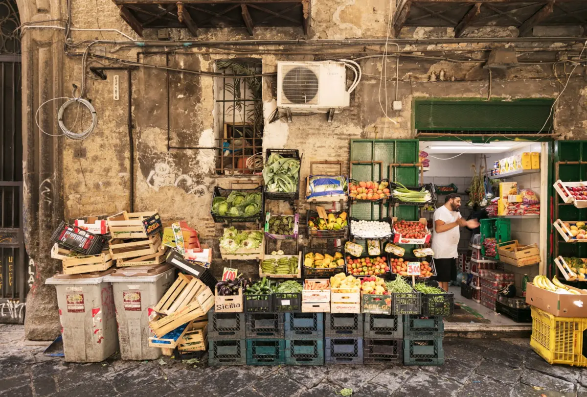 Market stall with crates of vegetables arranged outside a weathered stone wall; man in a white shirt stands among produce.