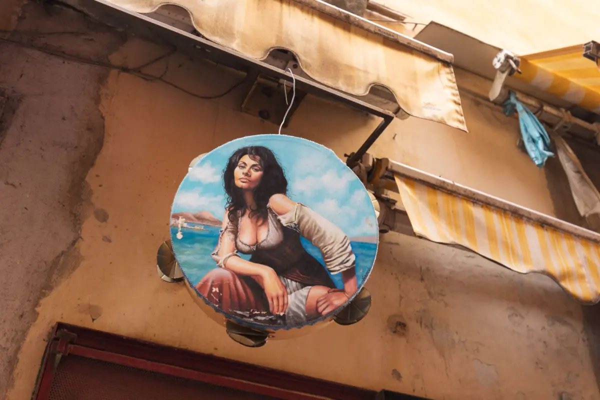 Circular painted sign of a dark-haired woman in a white blouse by the sea, hanging from a metal bracket under a striped yellow awning.