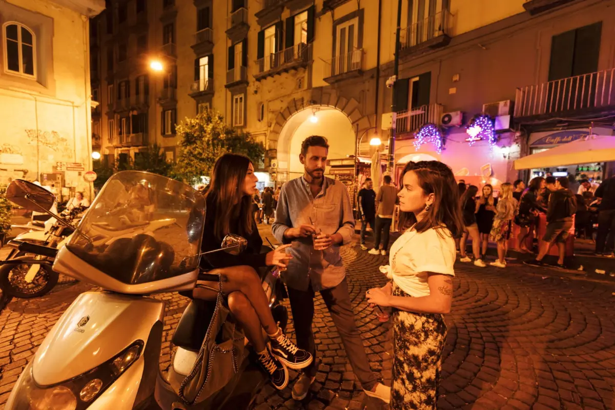 Nighttime street scene with a white motor scooter; three people chat on cobblestones, warm lights, arched entrance, crowd in background.
