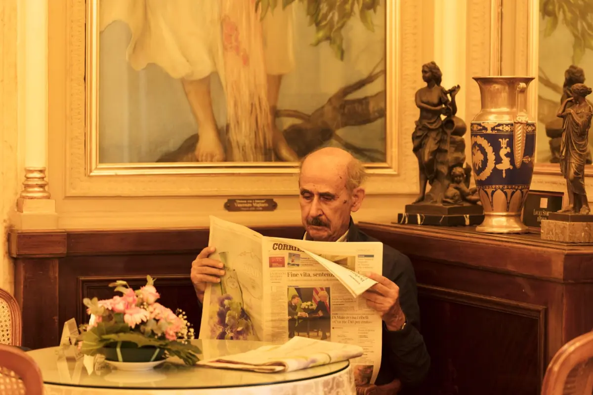 Older man reading a newspaper at a table in an ornate room with statues and a decorative vase.
