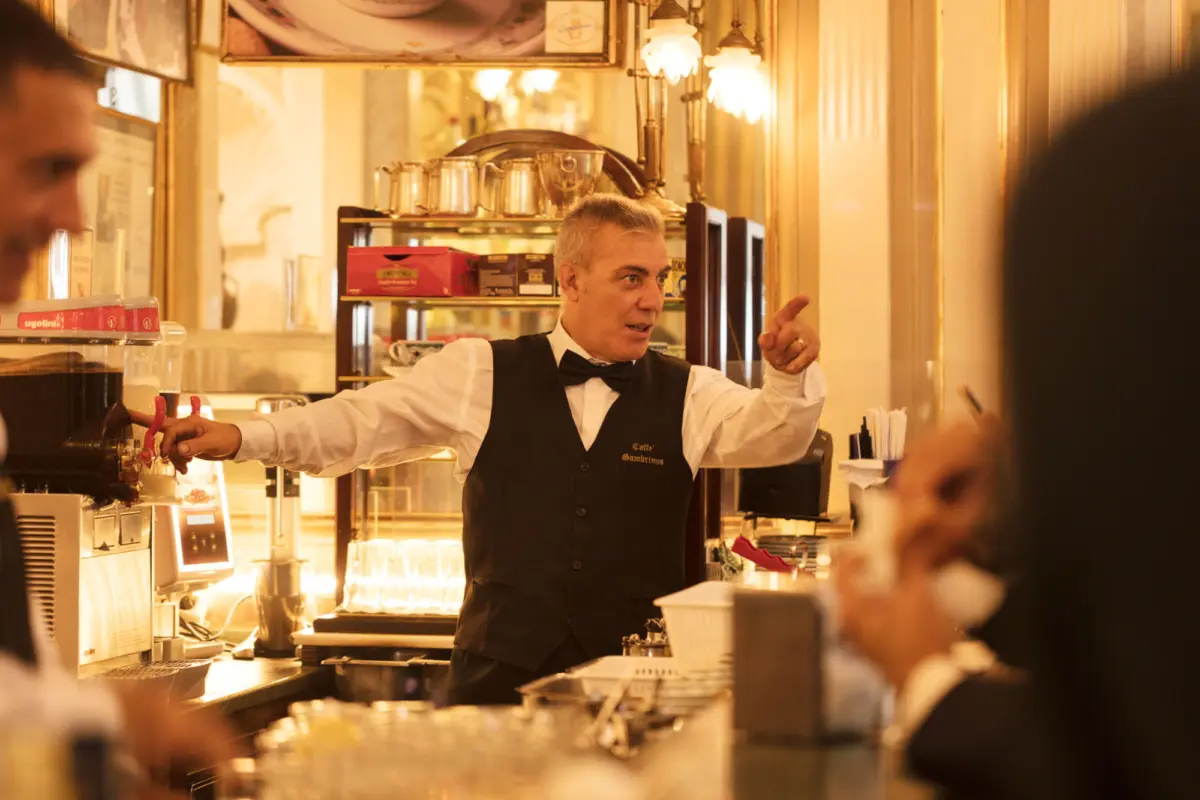 Bartender in a black waistcoat and bow tie points at customers behind a warmly lit cafe counter.