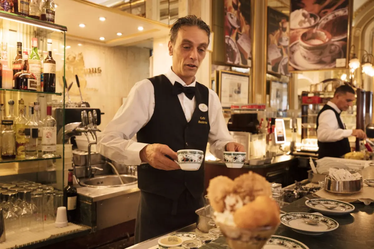 Server in black vest and bow tie balances floral teacups on a tray at a cafe bar; liquor bottles on glass shelves behind.