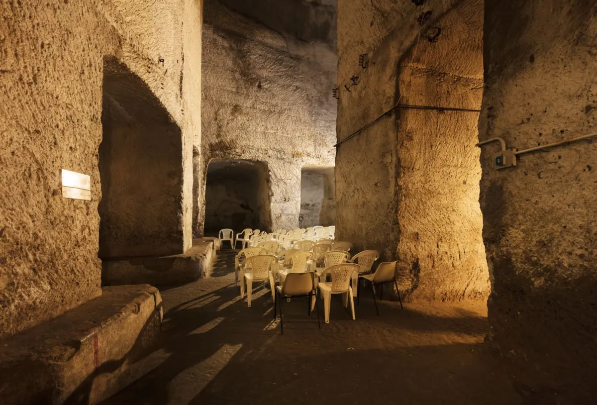 Stone-carved amphitheater with rows of beige plastic chairs set for an audience; rough cavern walls, arches, and warm lighting.