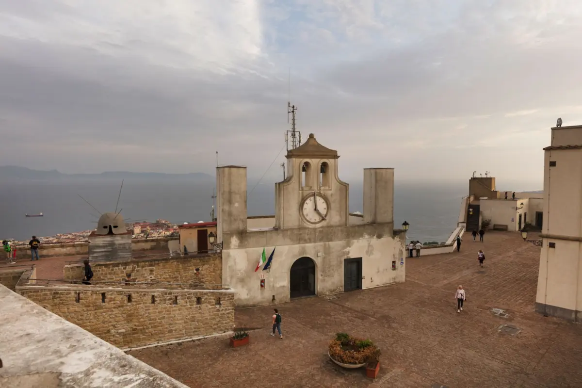Clock tower on hilltop plaza; pedestrians walk on cobblestone square overlooking the sea.