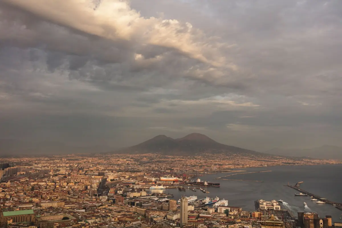 Aerial view of a busy harbor city with docks, ships, dense shoreline buildings, and distant volcanic mountains under a cloudy sky.