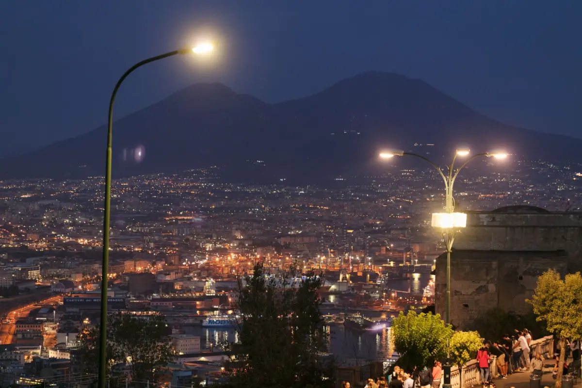 Night cityscape with distant mountain, curved streetlamps lit, and people strolling along a waterfront promenade.
