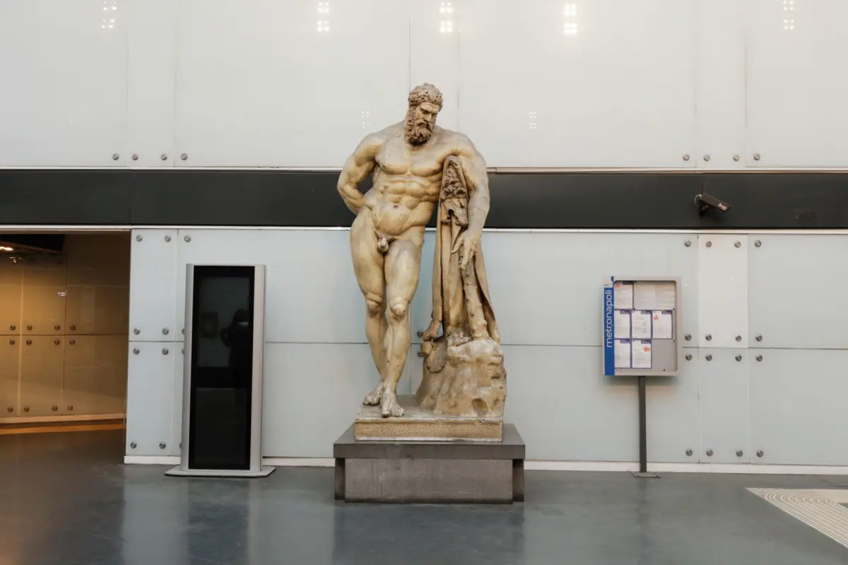 Stone statue of a nude, muscular bearded figure holding a draped cloak, set before a modern metal-paneled building.
