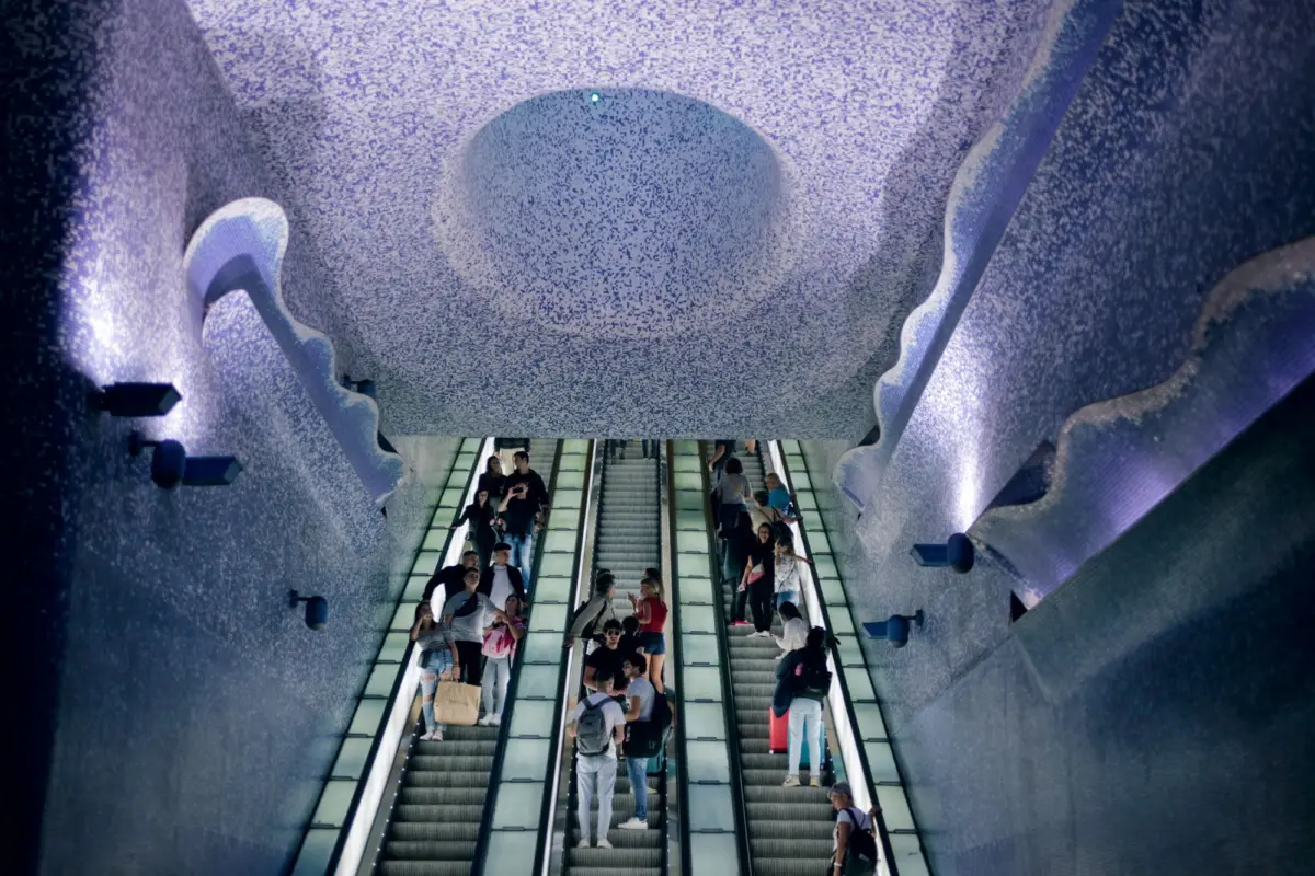 Escalators in a blue mosaic tunnel; crowds of pedestrians ride upward and downward.