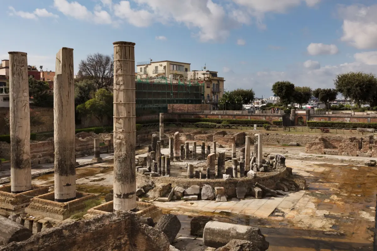 Ancient stone columns rise from a ruined harbor site, with modern buildings and trees along a waterfront promenade.