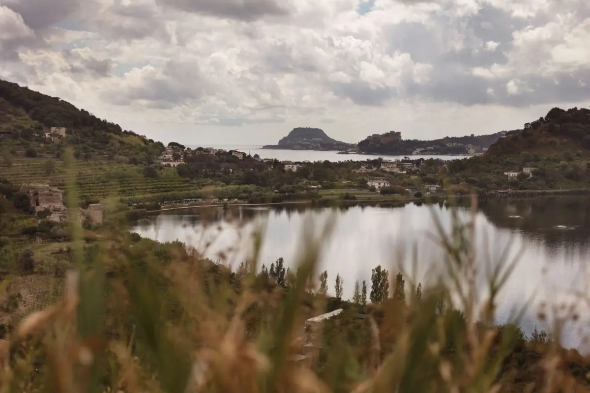 Panoramic lake valley with terraced fields, a small coastal town, and a distant fortress on a rocky island under cloudy skies.