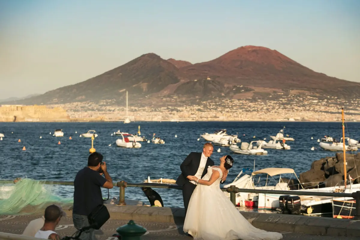 Wedding couple in a dress and suit dip on a harbor promenade as a photographer shoots, with boats and a mountain behind.