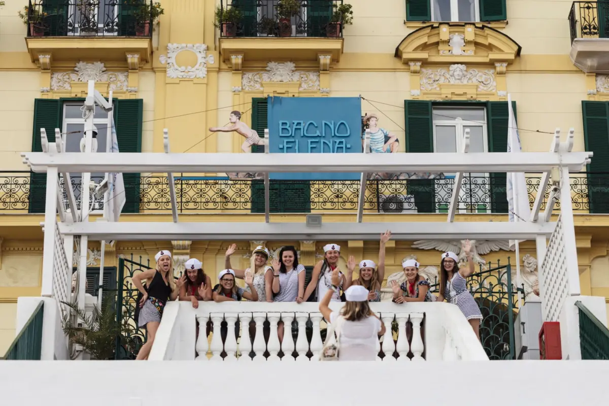Women in striped sailor outfits and hats wave from a yellow, ornate building balcony during a parade; white railing in foreground.
