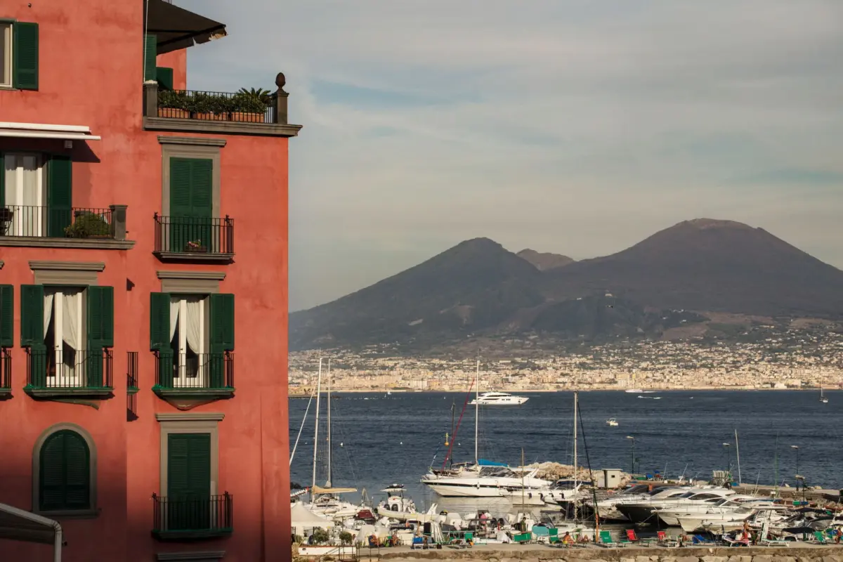Pink stucco apartment building with green shutters and balconies overlooking a marina; sailboats and distant mountains in the background.
