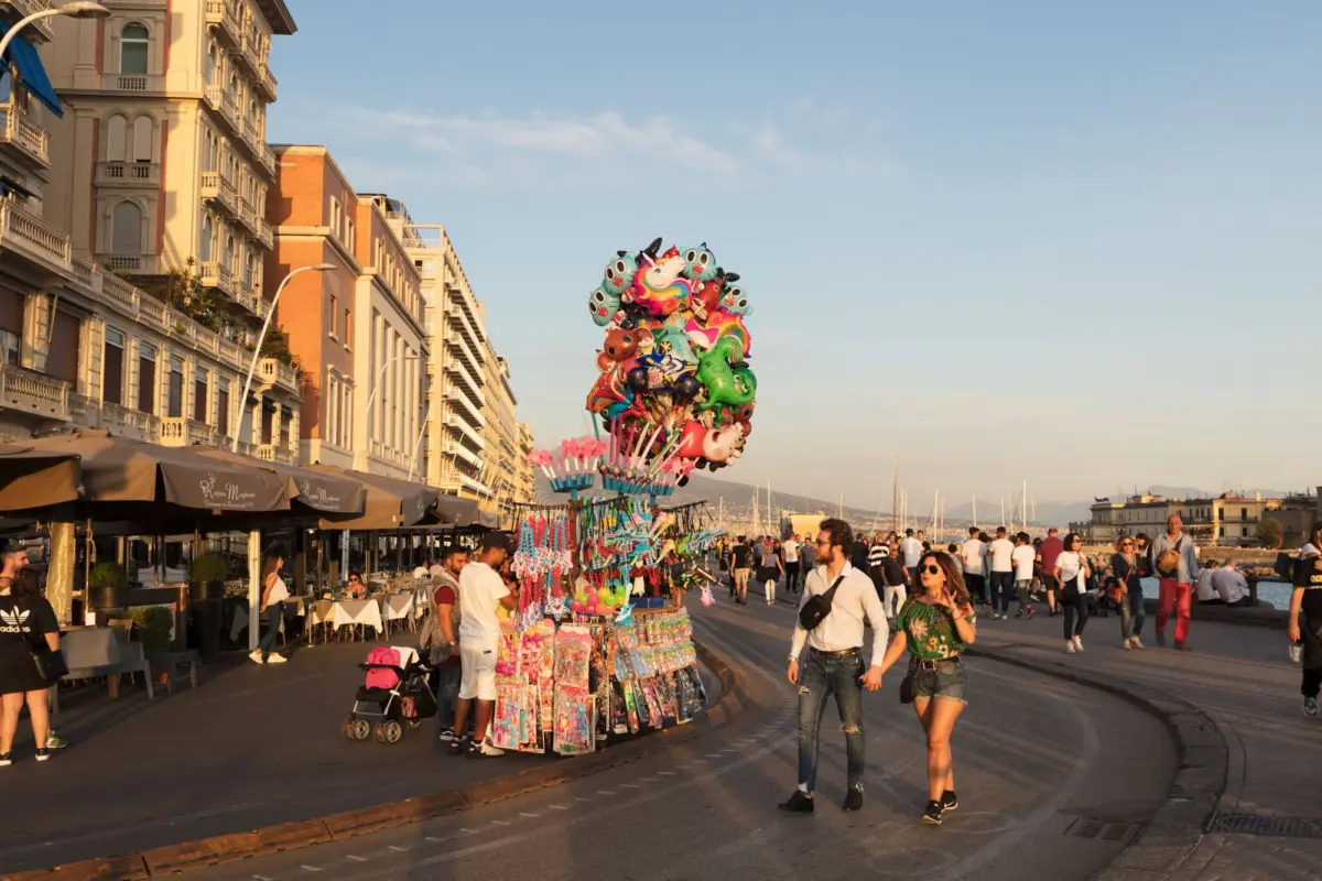 Colorful balloon vendor cluster on a seaside promenade; pedestrians stroll past historic buildings at sunset.