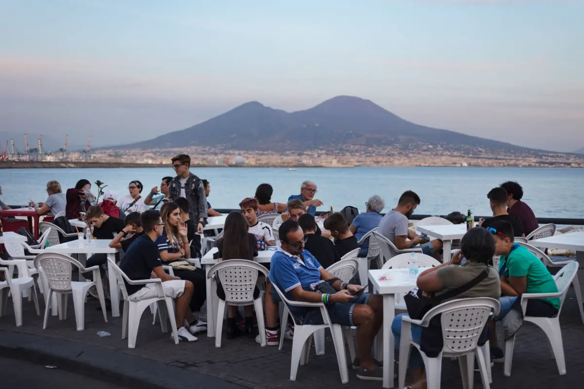 Group of diners sitting at white plastic tables along a seaside promenade, with a distant mountain and harbor in the background.