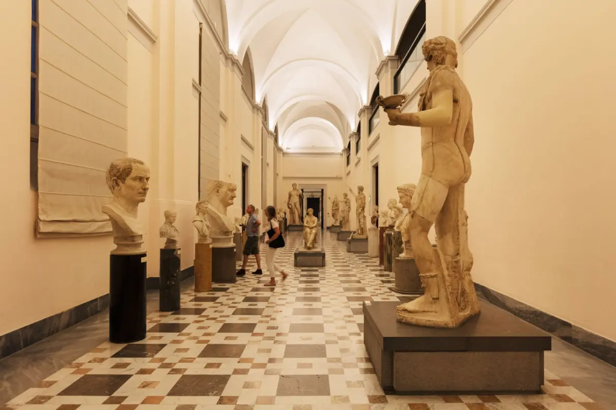 Museum gallery corridor lined with marble statues and busts; visitors walk the checkered floor beneath vaulted arches.