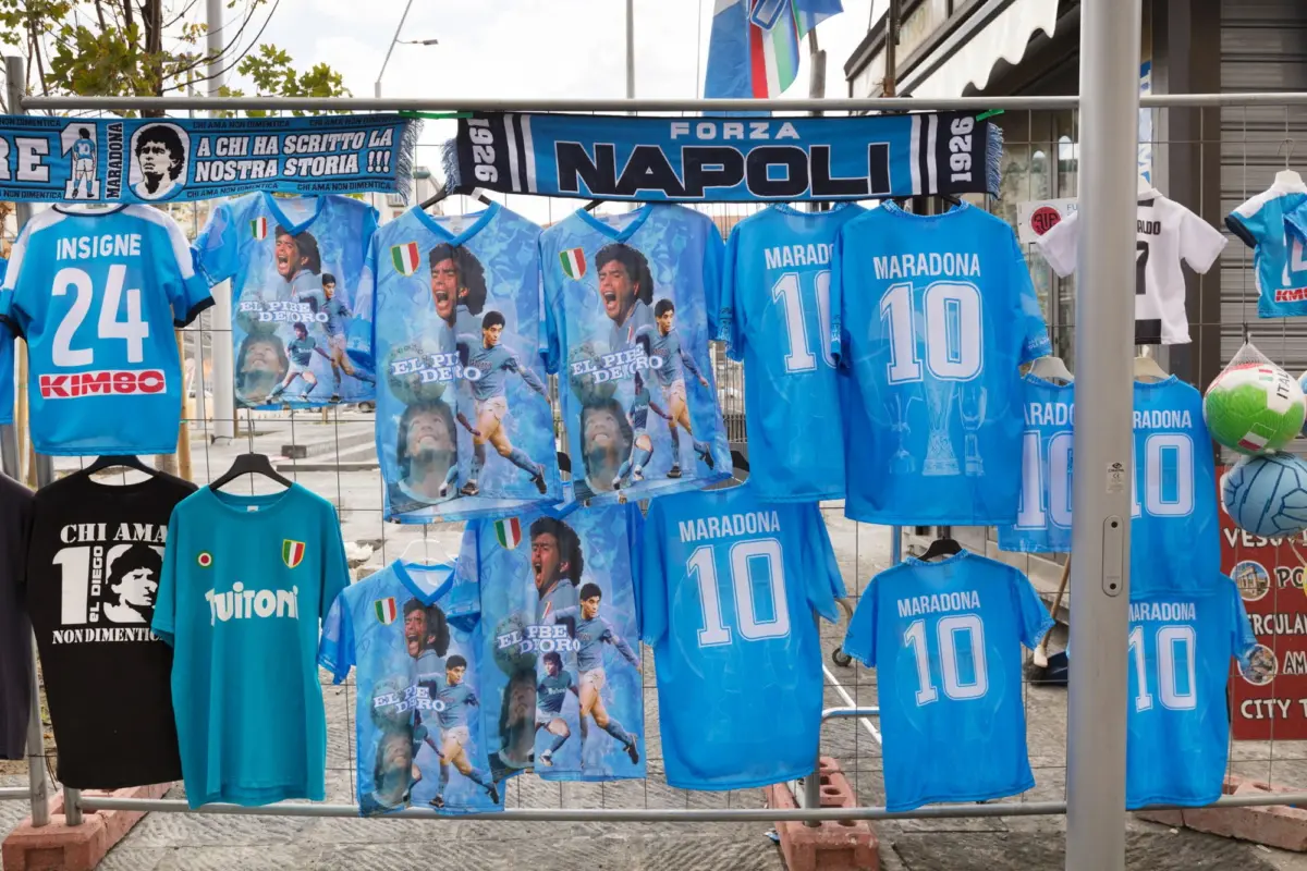 Blue Napoli football jerseys with Maradona imagery and MARADONA 10, hanging on display at an outdoor street market.