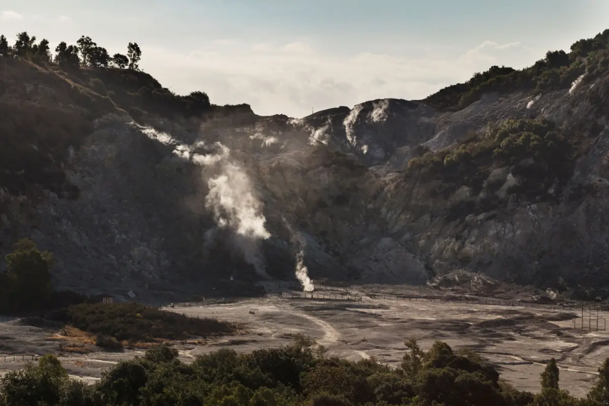 Geothermal volcanic crater with rising steam vents, rocky slopes, and sparse fencing, distant hills with trees under cloudy sky.