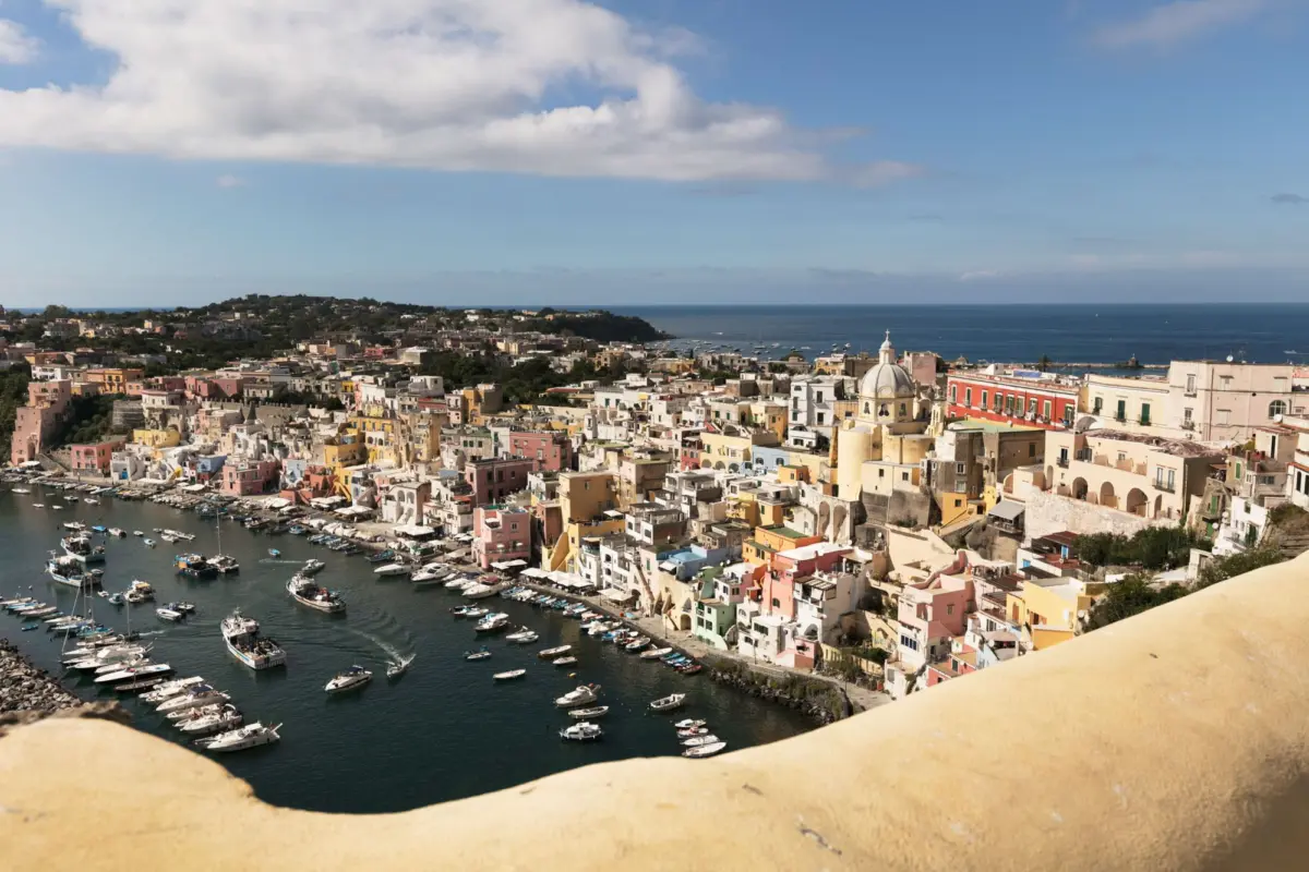 Pastel-colored Mediterranean harbor town with moored boats along the quay and blue sea beyond.