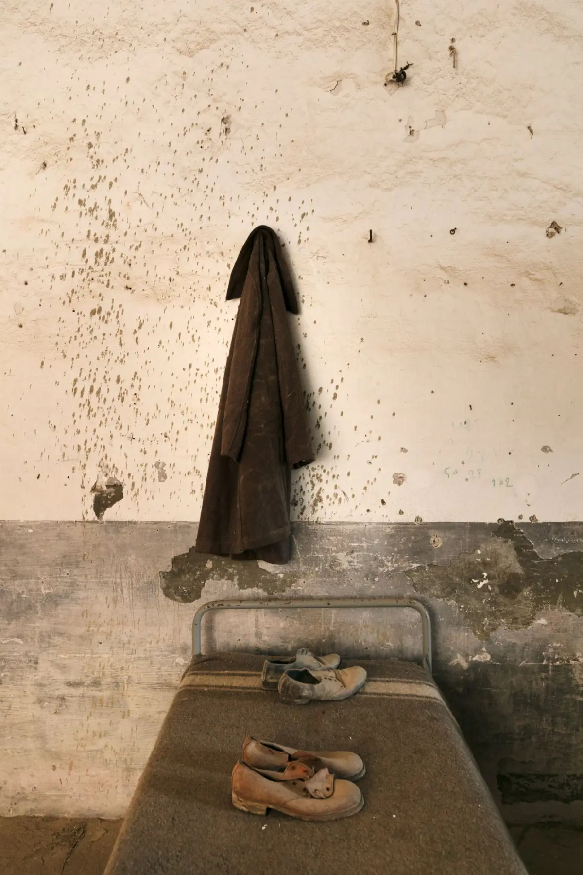 Brown overcoat hung on wall hook above a dusty bed with worn shoes in a dilapidated room.