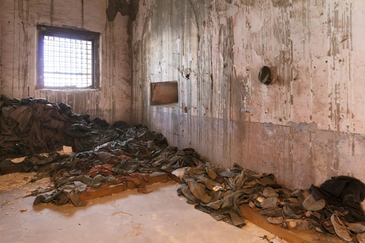 Abandoned room with peeling, stained walls, a barred window, and heaps of dark fabric scattered across the floor.