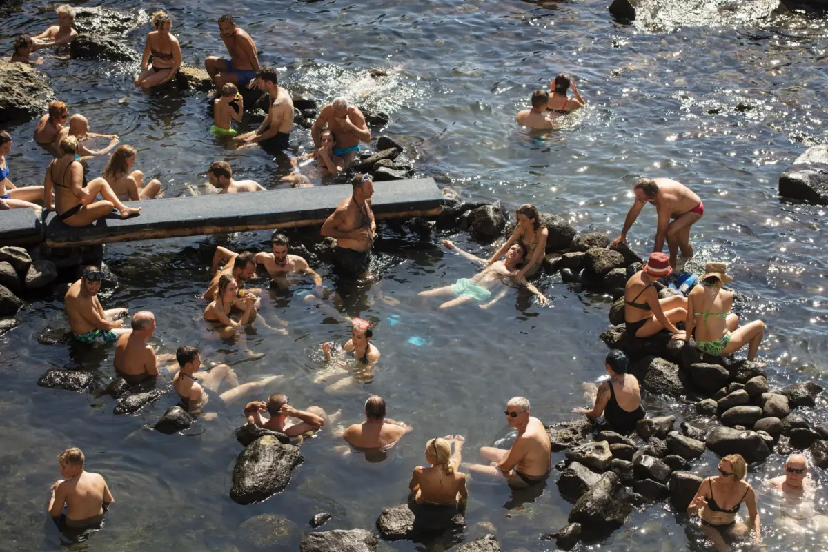 Crowded rocky coastline with many adults swimming, lounging on rocks and a wooden platform in sunlit blue water.