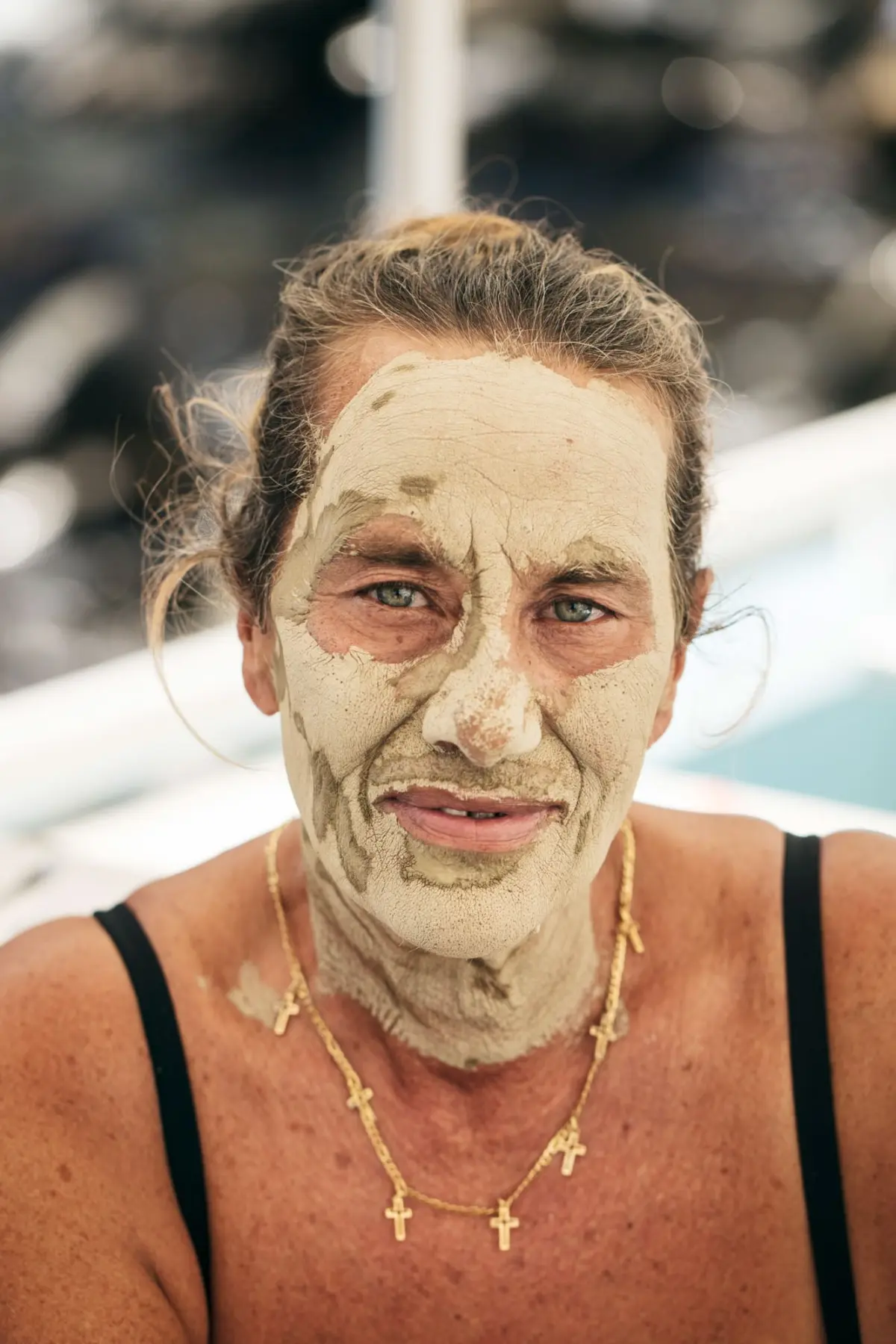Person with beige clay facial mask, looking at camera, wearing black strap top and gold cross necklaces, poolside outdoors.