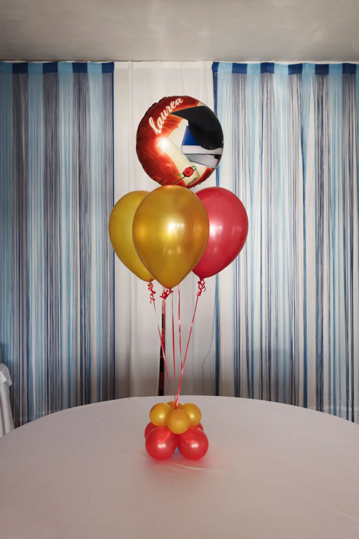 Balloon bouquet centerpiece on a round white table with red foil balloon atop yellow and pink balloons, blue striped backdrop.