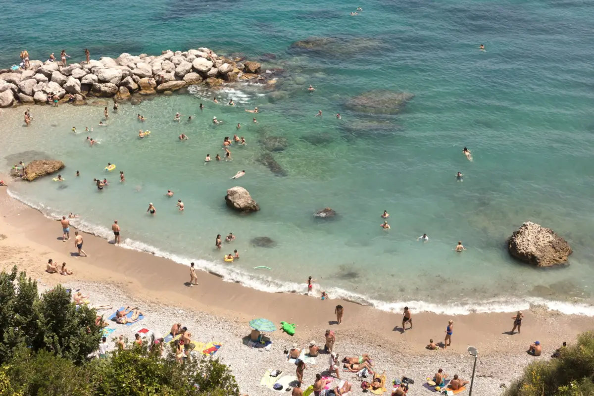 Crowded pebble beach with swimmers near a rocky breakwater and turquoise sea.