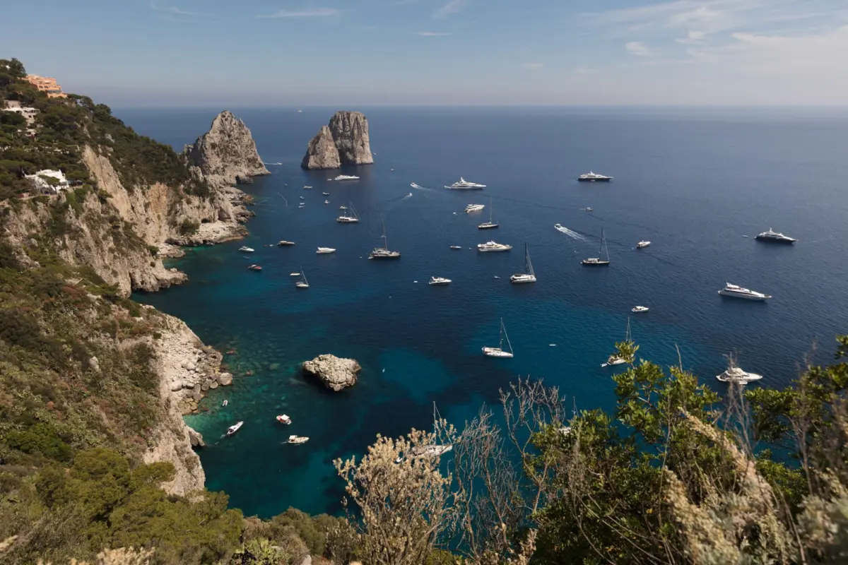 Cliffside houses on a rugged coastline overlook a blue sea; numerous yachts anchor nearby, with two rocky sea stacks in the distance.