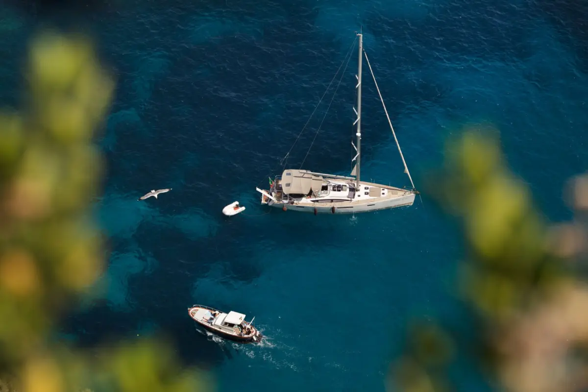 An aerial view of a sailing yacht anchored in turquoise sea, with two small boats nearby; blurred greenery in foreground.