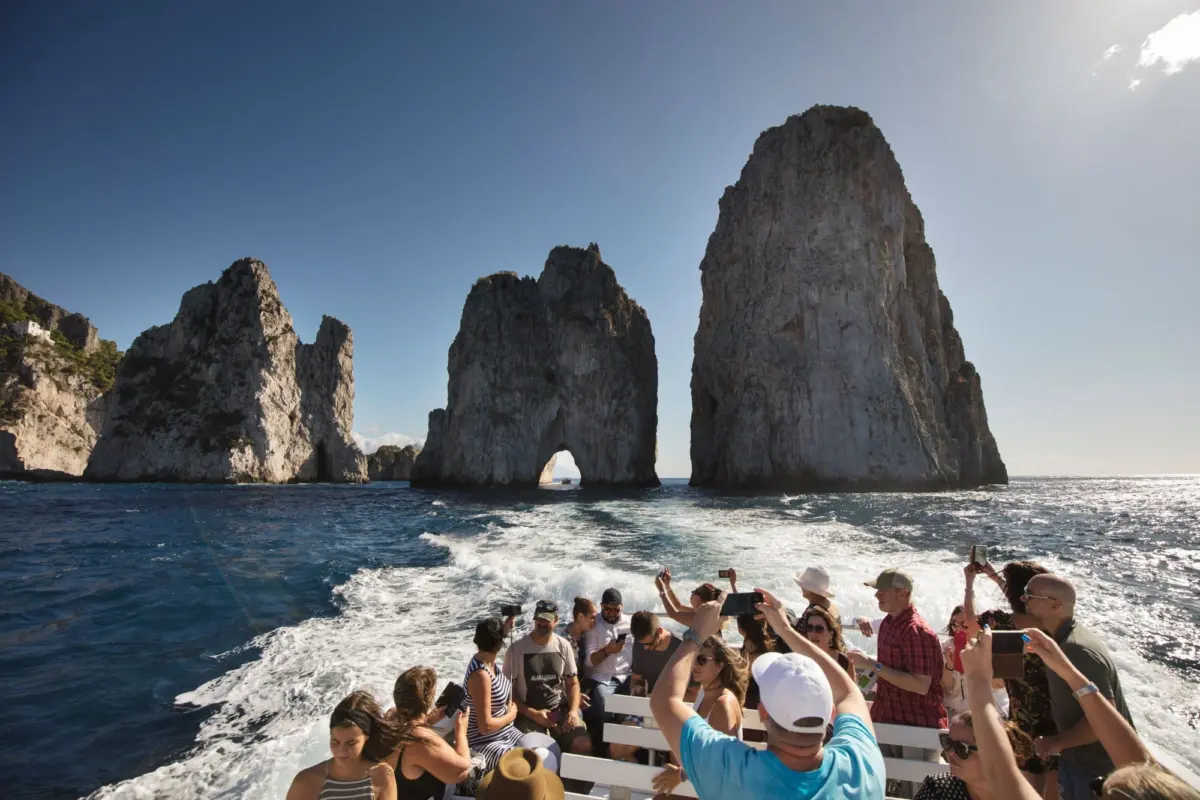 Group of tourists on a boat taking photos of towering sea stacks with an arch off a rocky coast.
