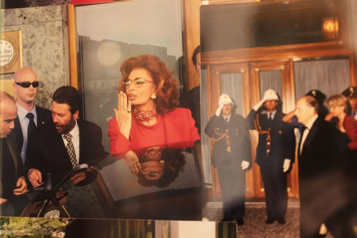 Red-haired person in red suit and sunglasses gesturing toward mouth, standing in front of a glass storefront with suited men and uniformed guards.
