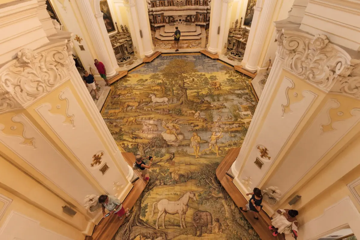 Ornate Baroque hall with tall white columns and a large animal mosaic floor; visitors stroll and observe.