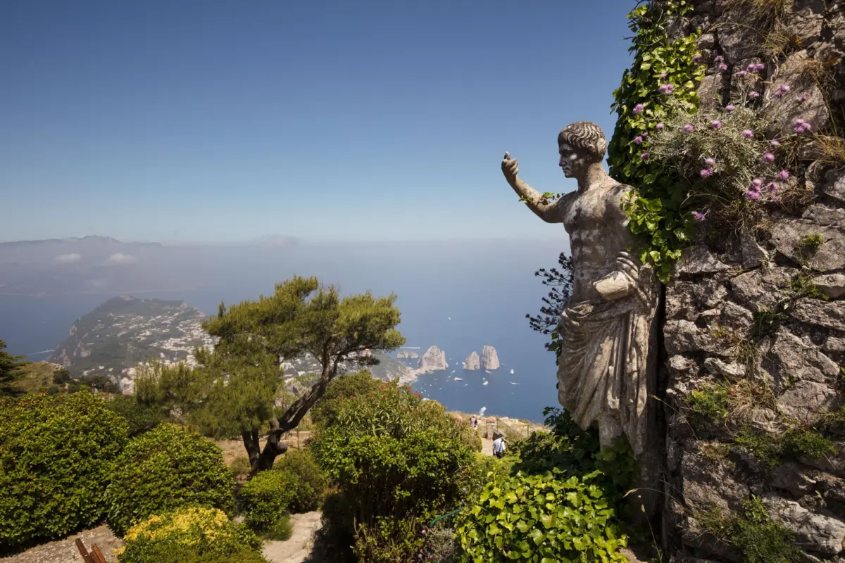 Stone statue of a man with outstretched arm on a rocky cliff, ivy and purple flowers surround, overlooking the sea and distant islands.