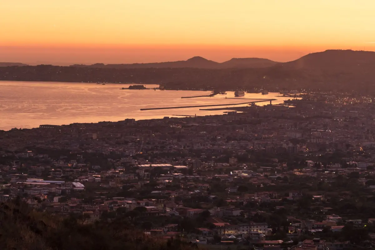 Coastal city skyline at sunset over a bay with harbor piers and distant hills bathed in orange light.