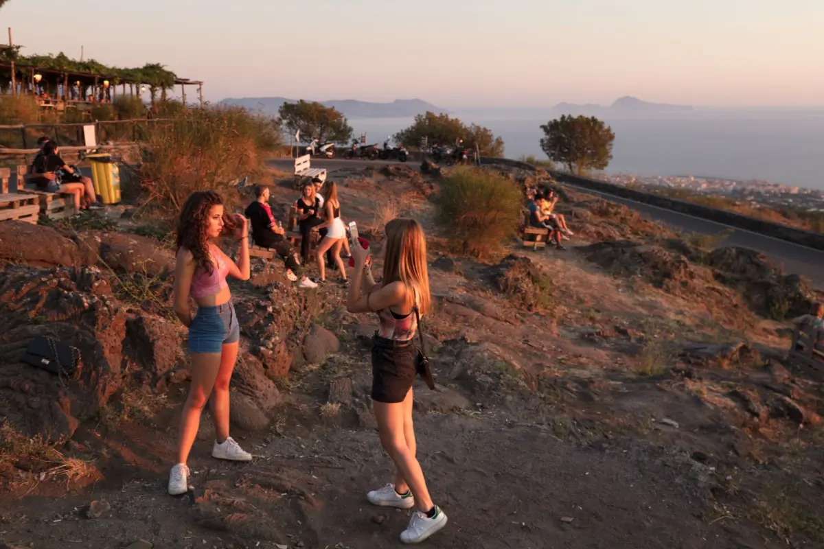 Group of young adults at a rocky seaside lookout at sunset, posing for photos with sea and distant islands in the background.