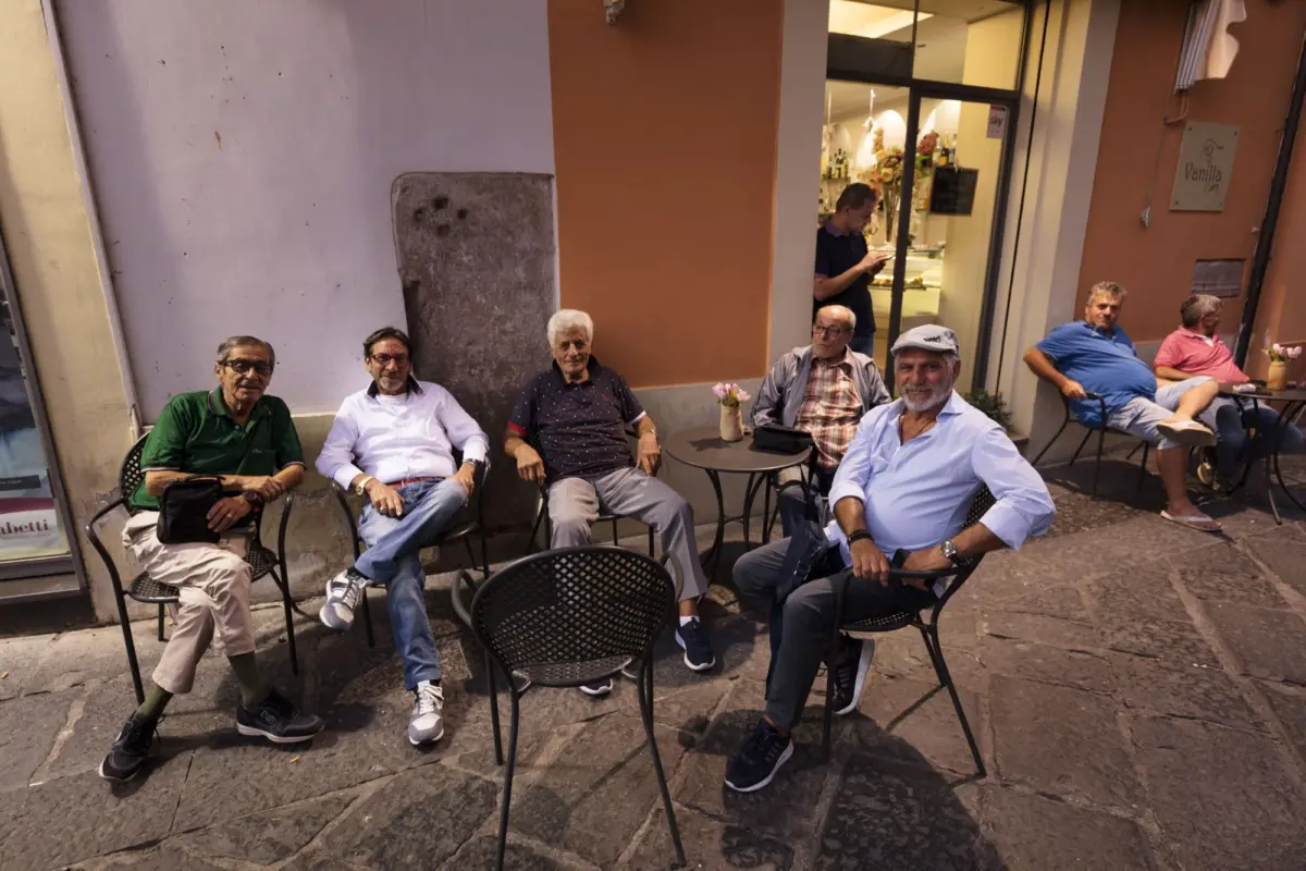 Group of elderly men seated at outdoor cafe tables on a city sidewalk; one man at the entrance uses a smartphone.