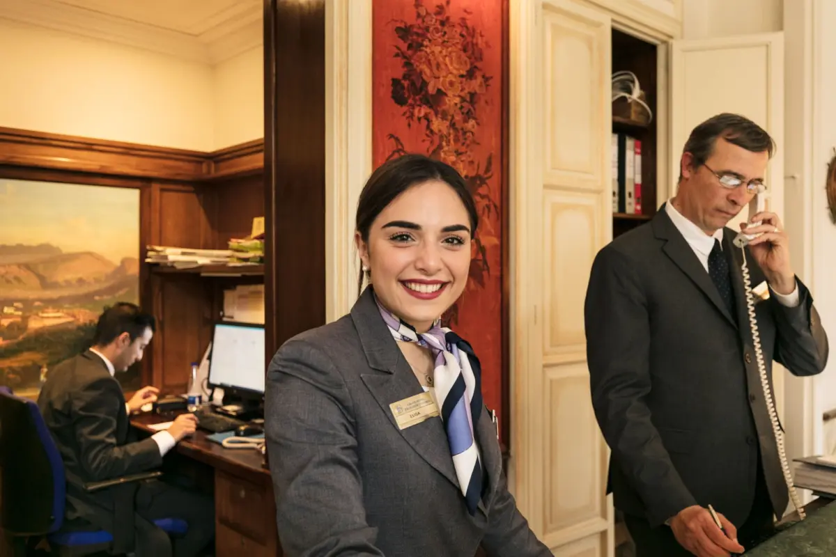 Smiling female receptionist in gray suit and scarf at a wood-paneled office reception; man in suit on phone in background.