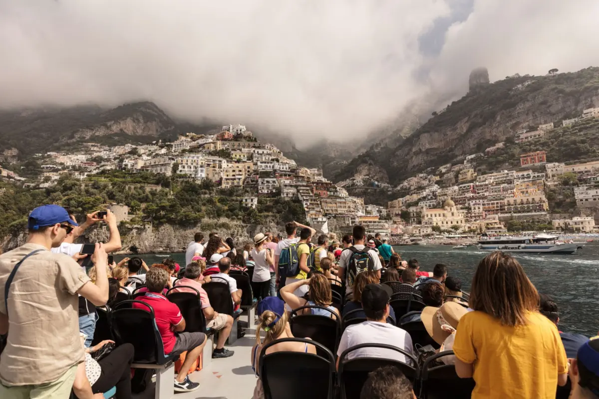 Tourists on an open-top boat photograph the Positano coast, with colorful hillside houses and misty mountains.
