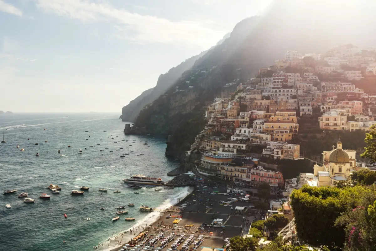 Steep hillside town of pastel buildings along a Mediterranean coast, with a busy beach, harbor, and numerous small boats.