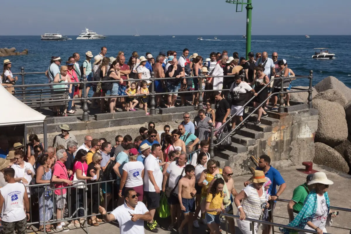 Crowd of tourists along a seaside promenade, many wearing hats, leaning on railing, with boats on the blue ocean behind.