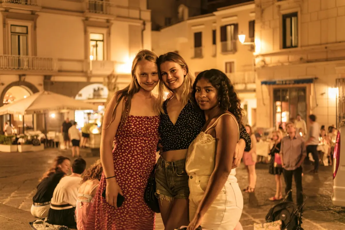 Three friends posing for a photo in a nighttime city square, with warmly lit buildings and outdoor cafe tables.