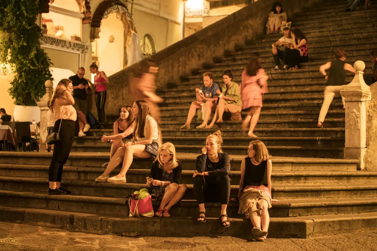Group of people seated and standing on a broad stone staircase in a nighttime plaza, with restaurant facades and arches in the background.