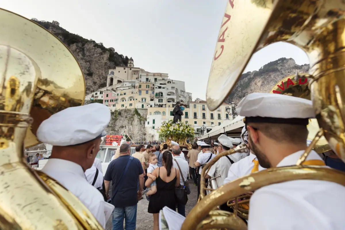 Brass band musicians aboard a boat perform in a crowded harbor beside pastel hillside townhouses.