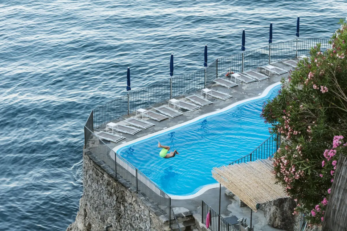 Person in lime-green swimsuit dives into a curved blue pool beside a seaside terrace with lounge chairs, closed blue umbrellas, and flowering shrubs.