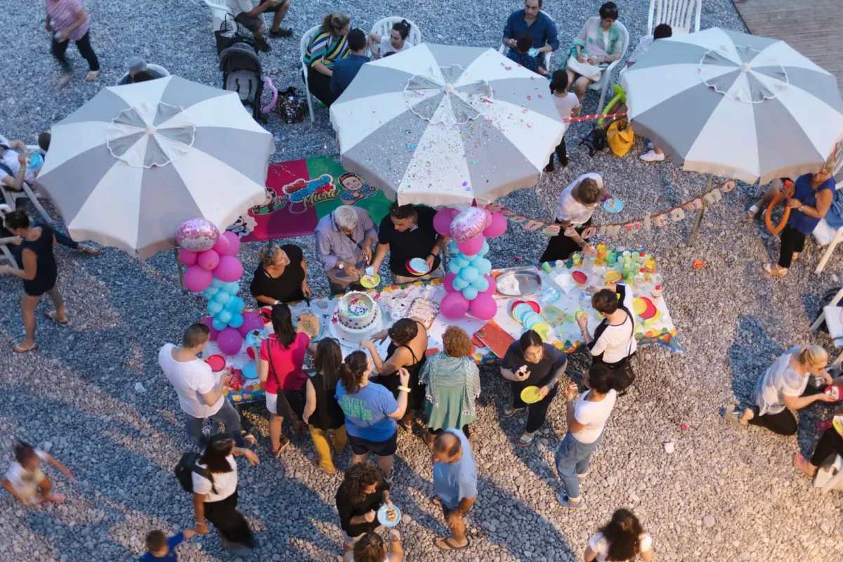 Guests gather around a decorated table with cake at an outdoor gravel venue under striped umbrellas.
